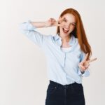 Beautiful redhead woman showing peace or victory signs and looking sassy at camera, standing against white background.